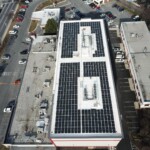Aerial view of the storage facility that shows the building with the indoor units and offices, the parking lot, and surroundin roadways. On the rood of the building is covered in solar panels