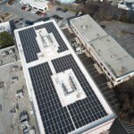 Aerial view of the storage facility that shows the building with the indoor units and offices, the parking lot, and surroundin roadways. On the rood of the building is covered in solar panels