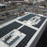Aerial view of the storage facility that shows the building with the indoor units and offices, the parking lot, and surroundin roadways. On the rood of the building is covered in solar panels