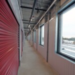Hallway with the left side of the hall lined by storage units with red roll up and doors and the right side with a large window that lets in a lot of light and overlooks the surrounding area (a roadway)
